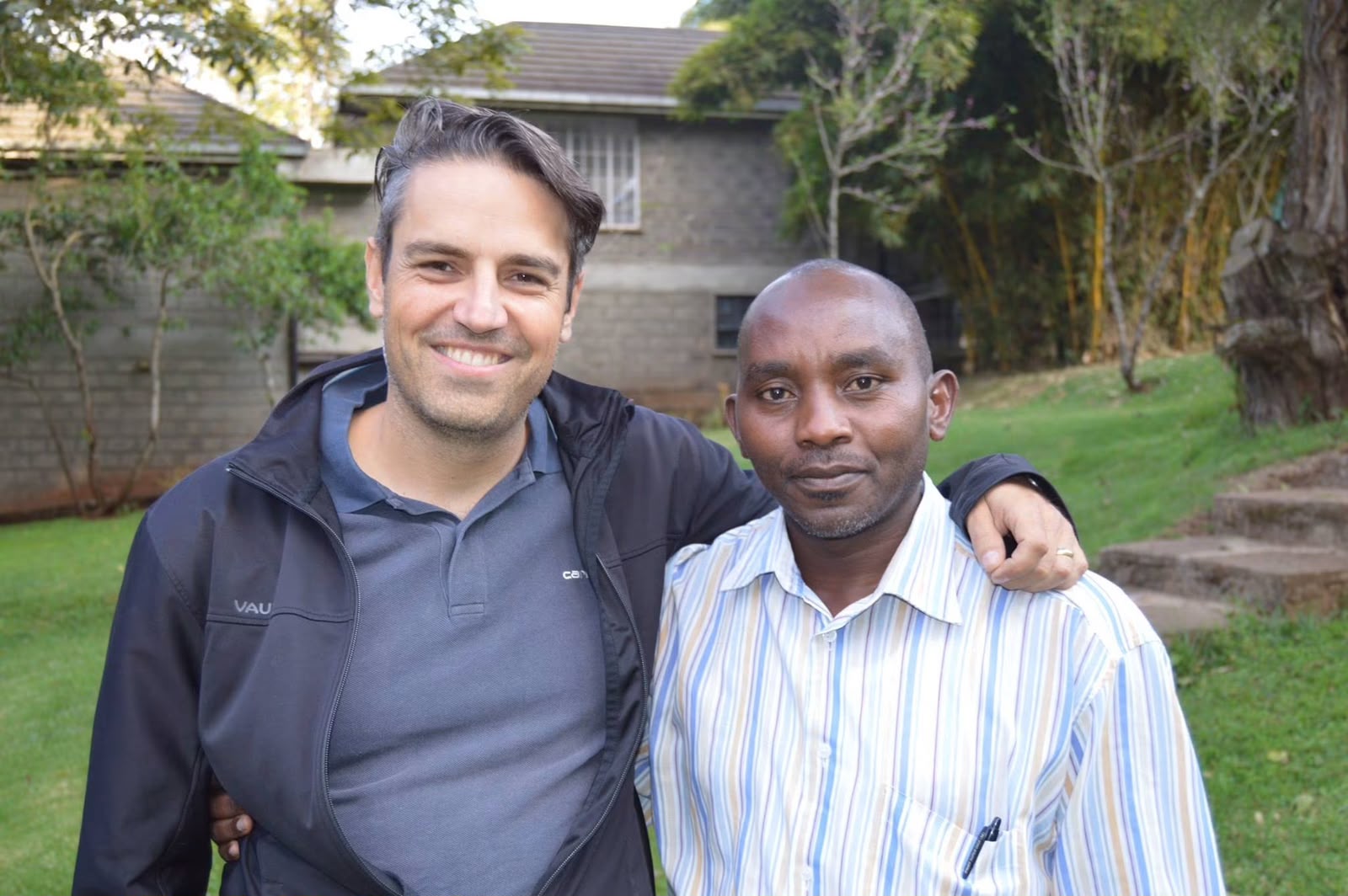Two founding members smile together outdoors in the gardens at Thayu resort in Limuru, one with his arm around the other's shoulder, during the September 2016 SPEC founding retreat.